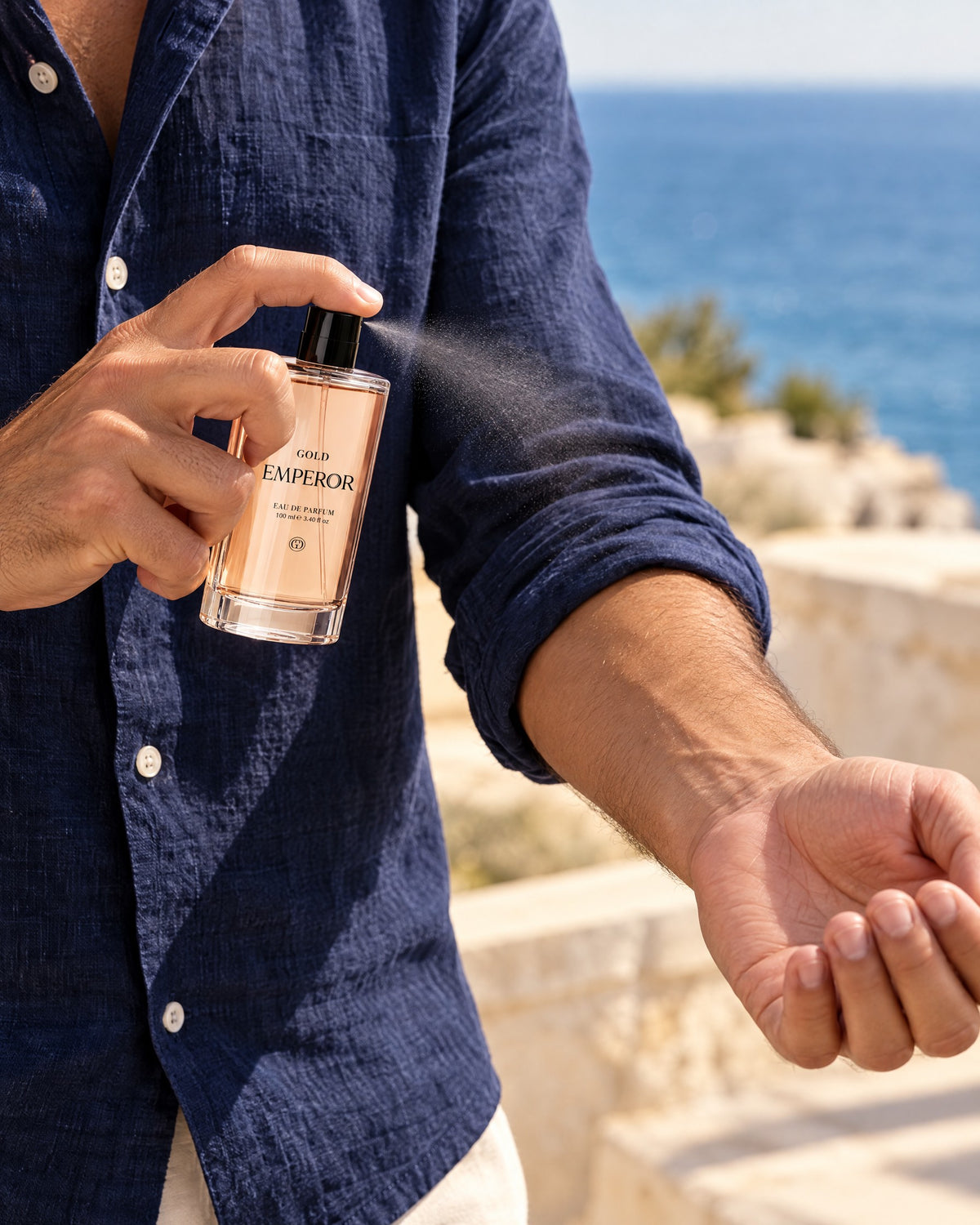 Mann im blauen Leinenhemd sprüht GOLD Emperor Eau de Parfum am Meer, holzig-frischer Herrenduft