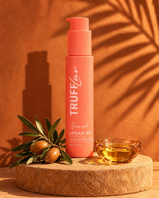 TRUFFLuv Argan Oil Haaröl in korallfarbener Flasche auf Sandstein neben Arganzweig und Schale mit Arganöl